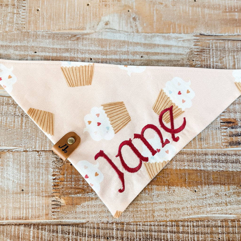 Dog bandana with 'Jane' printed on it, featuring cupcake designs, on a wooden surface.