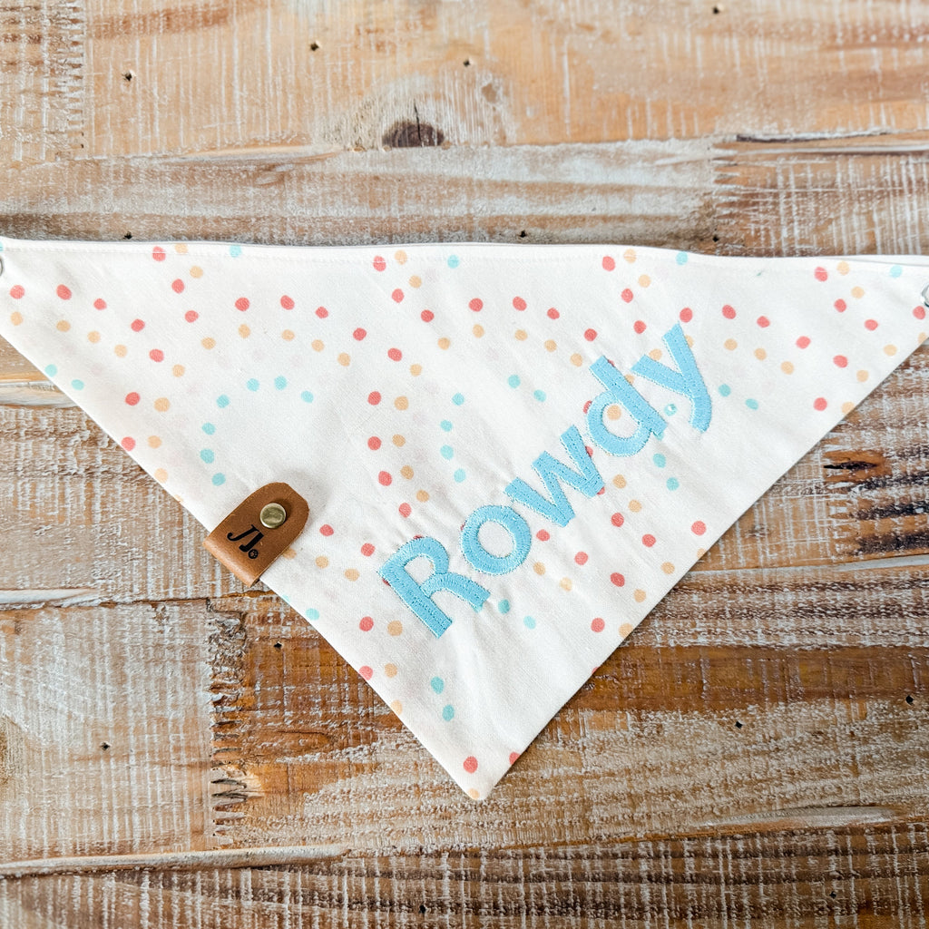Dog bandana with 'Rowdy' printed on it, featuring colorful polka dots, on a wooden surface.